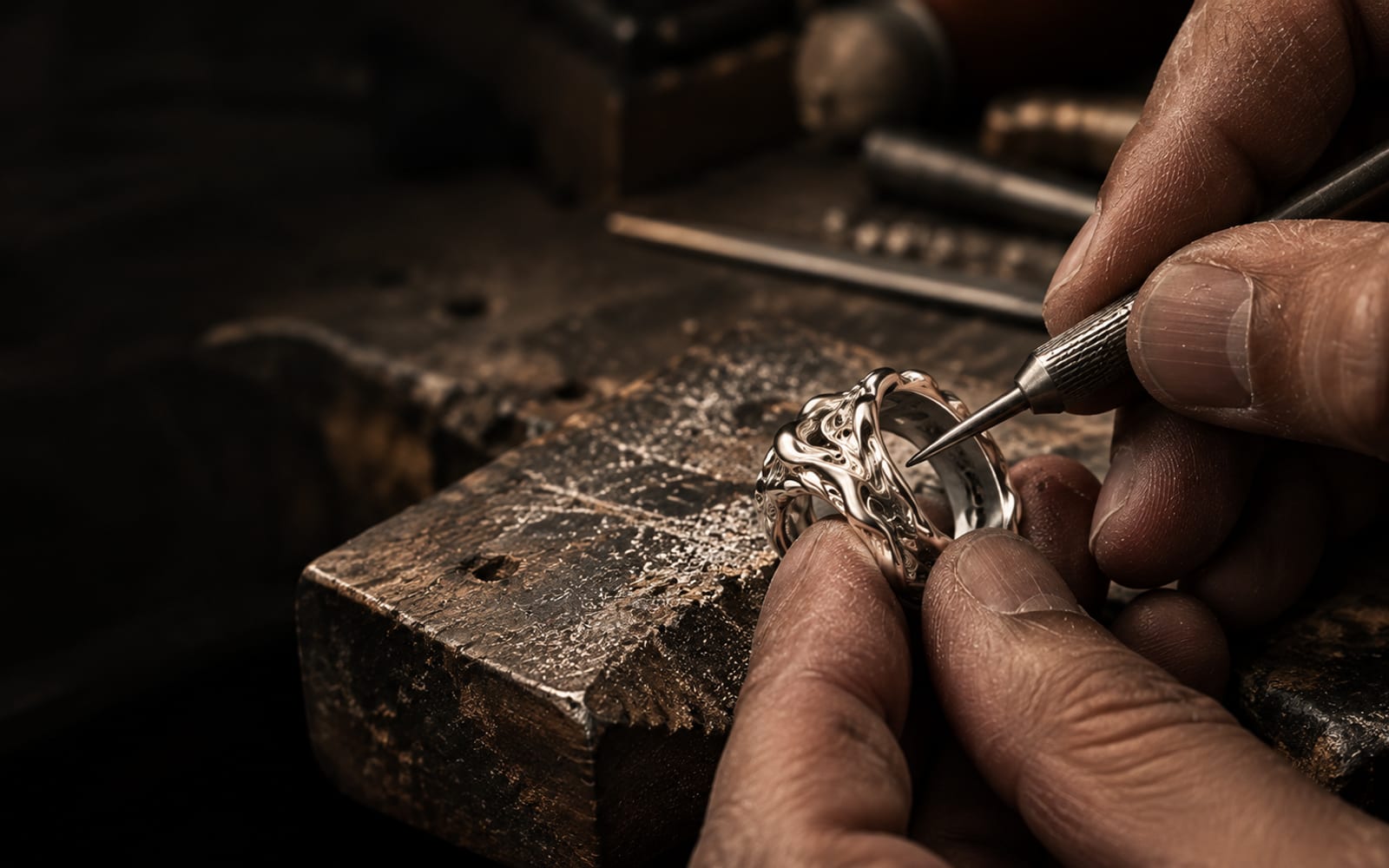 A jeweller’s hands working a silver ring at the bench under a single light.