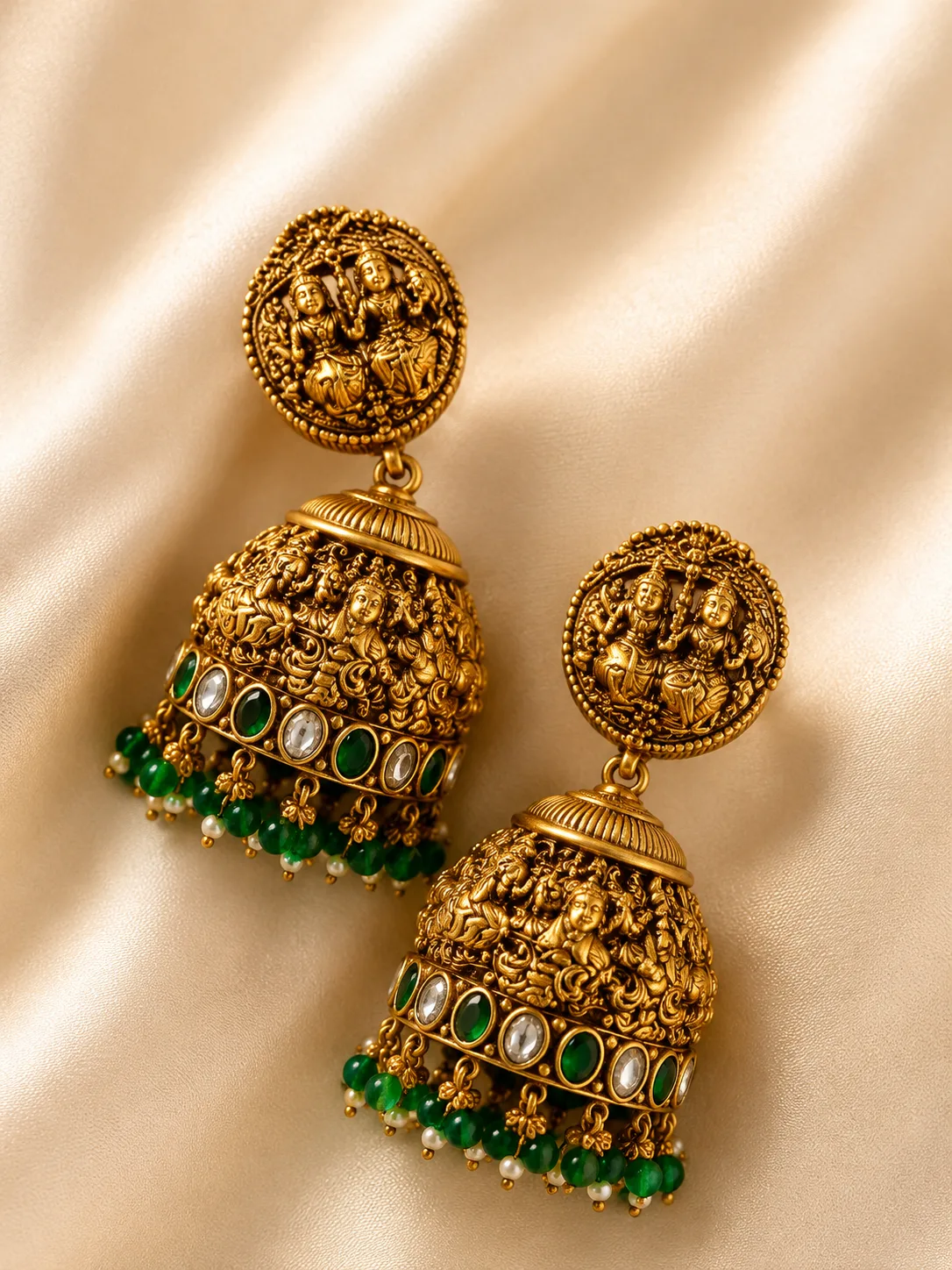Pair of gold-tone dome-shaped drop earrings with figural relief detail, a band of green and colourless stones, and a fringe of green and white beads, resting on cream satin.