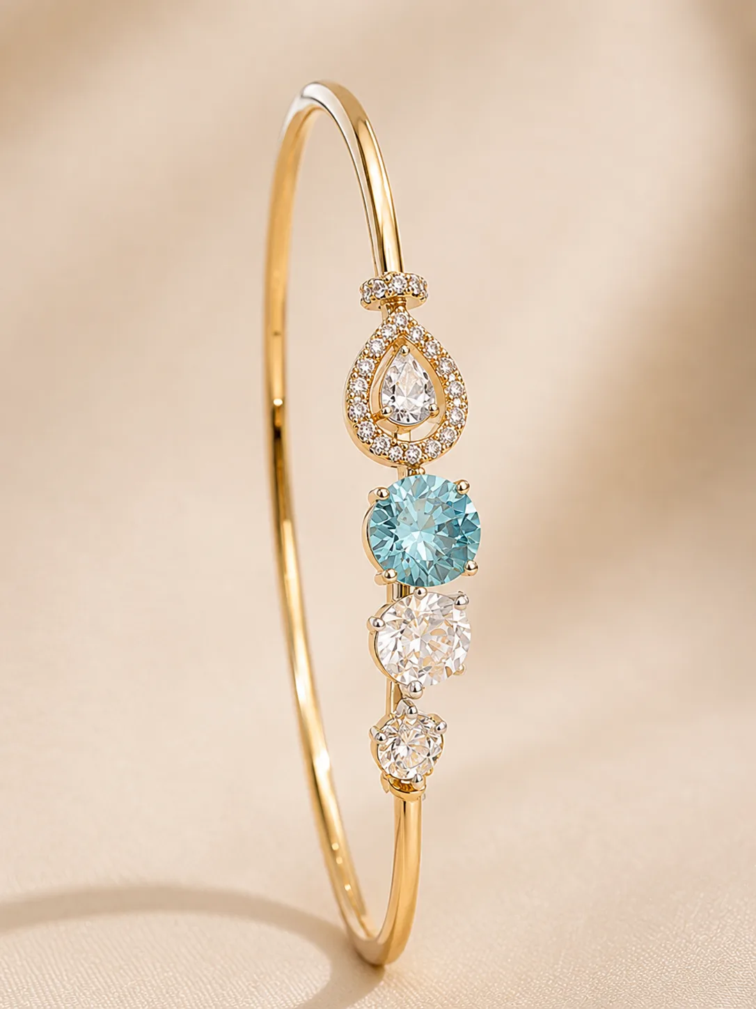 A fine gold-tone open cuff set with a pear-cut colourless stone in a halo, a round blue stone and smaller colourless stones, on cream satin.