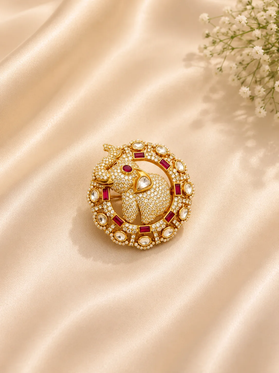 A round gold-tone jewel with an elephant motif set in small colourless stones, framed by red rectangular and flat colourless stones, lying on cream satin beside white flowers.