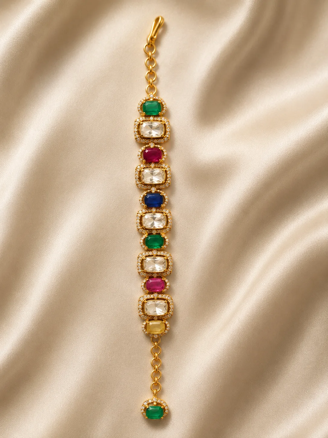 A flat gold-tone bracelet lying on cream satin, set with a single row of green, pink, blue, yellow and colourless rectangular stones, each framed by small colourless stones, with an adjustable chain, a lobster clasp at one end and a green oval stone drop at the other.