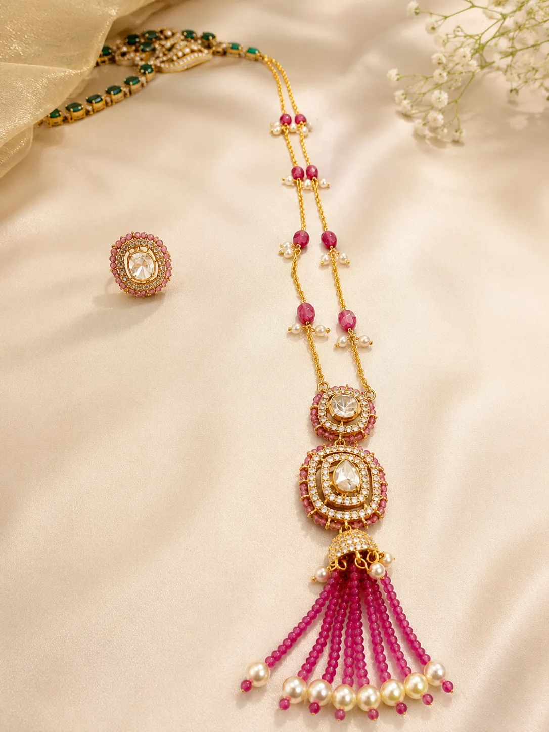 A long double gold-tone chain set with pink beads and small white beads, ending in two round medallions with large colourless stones and a tassel of pink beaded strands tipped with white drops, laid on cream satin beside a matching round ring; a green-stone necklace lies partly in frame at the top left.
