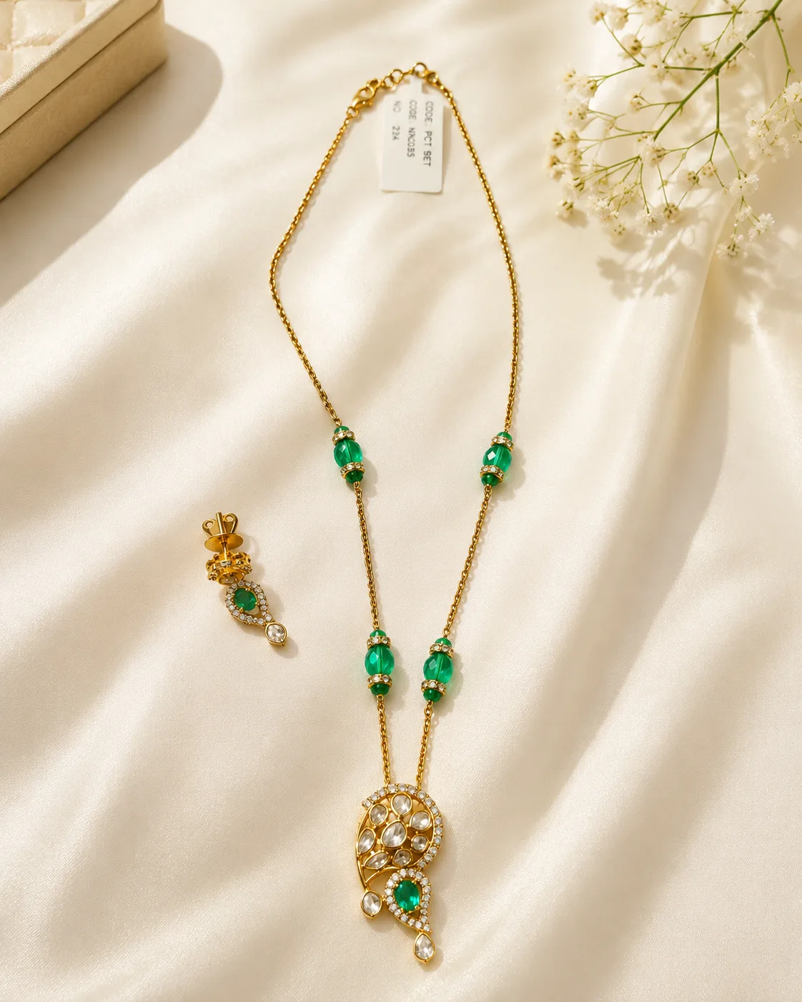 Gold-tone chain necklace with green oval stones spaced along the chain, ending in a round pendant of small colourless stones with a green oval stone below, shown with one matching drop earring on cream fabric beside white flowers.